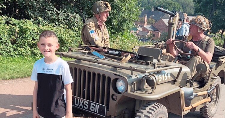 little boy stand and smiles next to historic army vehicle with two costumed men